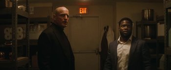 Movie still from “The Man from Toronto” (2022), directed by Patrick Hughes – Two men standing next to each other in front of an exit sign; Medium shot, Over the shoulder angle
