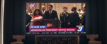 Movie still from “The Man from Toronto” (2022), directed by Patrick Hughes – A television screen showing a news story about venezuelan president arvato speaking; Medium shot, Over the shoulder angle