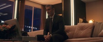 Movie still from “The Man from Toronto” (2022), directed by Patrick Hughes – A man in a suit sitting on top of a couch; Medium shot, Low angle
