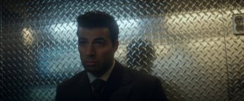 Movie still from “The Man from Toronto” (2022), directed by Patrick Hughes – A man wearing a suit and tie standing in front of a metal wall; Medium shot, Over the shoulder angle
