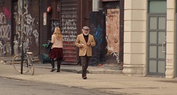 Movie still from “The Meyerowitz Stories” (2017), directed by Noah Baumbach – An older man walking down the street while a woman walks behind him; Wide shot, Over the shoulder angle