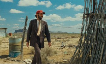 Movie still from “Paris, Texas” (1984), directed by Wim Wenders – A man wearing a suit and a baseball cap walking across a field; Wide shot, Low angle