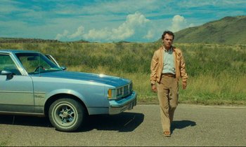 Movie still from “Paris, Texas” (1984), directed by Wim Wenders – A man walking next to a blue car on a road; Wide shot, Low angle