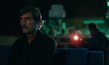 Movie still from “Paris, Texas” (1984), directed by Wim Wenders – A man sitting on a blue couch in a dark room; Close Up shot, Over the shoulder angle