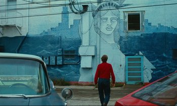 Movie still from “Paris, Texas” (1984), directed by Wim Wenders – A man in a red shirt is standing in front of a statue of liberty; Wide shot, Low angle