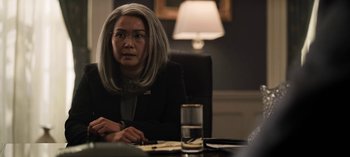 Movie still from “The Night Agent” (2023), created by Shawn Ryan – A woman sitting at a table in front of a glass of water; Medium shot, Over the shoulder angle