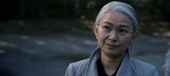 Movie still from “The Night Agent” (2023), created by Shawn Ryan – An older asian woman wearing glasses and a gray coat; Close Up shot, Over the shoulder angle