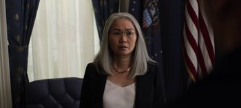 Movie still from “The Night Agent” (2023), created by Shawn Ryan – An older woman with long gray hair wearing a white blouse; Close Up shot, Over the shoulder angle