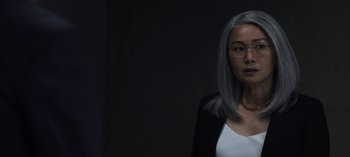 Movie still from “The Night Agent” (2023), created by Shawn Ryan – A woman with gray hair and glasses in front of a black background; Close Up shot, Over the shoulder angle