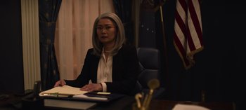 Movie still from “The Night Agent” (2023), created by Shawn Ryan – A woman sitting at a desk in front of an american flag; Medium shot, Over the shoulder angle
