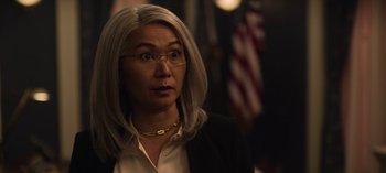 Movie still from “The Night Agent” (2023), created by Shawn Ryan – An older woman wearing glasses and a black jacket; Close Up shot, Over the shoulder angle