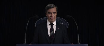 Movie still from “The Night Agent” (2023), created by Shawn Ryan – A man standing in front of microphones wearing a suit; Close Up shot, Low angle