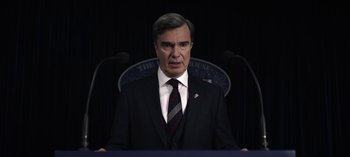 Movie still from “The Night Agent” (2023), created by Shawn Ryan – A man wearing a suit and tie standing in front of microphones; Close Up shot, Low angle