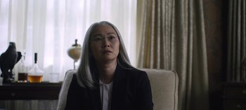 Movie still from “The Night Agent” (2023), created by Shawn Ryan – An older woman sitting on a couch in front of a window; Close Up shot, Over the shoulder angle