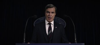 Movie still from “The Night Agent” (2023), created by Shawn Ryan – A man wearing a suit and tie standing in front of microphones; Close Up shot, Low angle