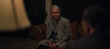 Movie still from “The Night Agent” (2023), created by Shawn Ryan – An older woman sitting on top of a brown leather couch; Medium shot, Over the shoulder angle