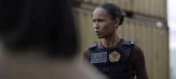 Movie still from “The Night Agent” (2023), created by Shawn Ryan – A woman in a black vest with a badge on her chest; Close Up shot, Over the shoulder angle