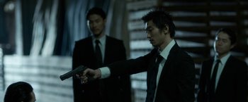 Movie still from “The Outsider” (2018), directed by Martin Zandvliet – A man in a suit and tie holding a gun in front of another man in a suit and tie; Medium shot, Over the shoulder angle
