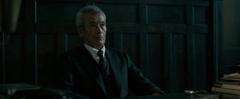 Movie still from “The Outsider” (2018), directed by Martin Zandvliet – An older man in a suit and tie sitting in front of a wooden wall; Close Up shot, Low angle