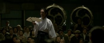 Movie still from “The Outsider” (2018), directed by Martin Zandvliet – A man holding a fan while standing in front of an audience; Medium shot, Low angle