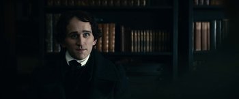 Movie still from “The Pale Blue Eye” (2022), directed by Scott Cooper – A man wearing a suit and tie standing in front of a book shelf; Close Up shot, Over the shoulder angle
