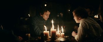 Movie still from “The Pale Blue Eye” (2022), directed by Scott Cooper – A man sitting in front of a group of lit candles; Medium shot, Over the shoulder angle