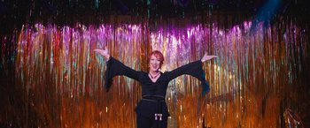 Movie still from “The Prom” (2020), directed by Ryan Murphy – A woman in a black outfit with her arms outstretched in front of a colorful background; Medium shot, Low angle