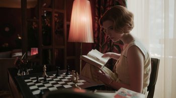 Movie still from “The Queen's Gambit” (2020), directed by Scott Frank – A woman reading a book while sitting at a chess board; Medium shot, High angle