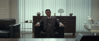 Movie still from “The Raid 2” (2014), directed by Gareth Evans – A man in a suit and tie sitting in a chair in a room; Medium shot, Low angle