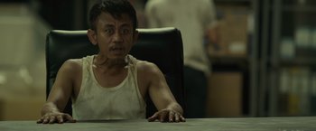 Movie still from “The Raid 2” (2014), directed by Gareth Evans – A man sitting at a table in front of an audience; Close Up shot, Over the shoulder angle