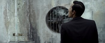Movie still from “The Raid 2” (2014), directed by Gareth Evans – A man in a black suit blowing smoke out of his mouth; Close Up shot, Low angle