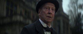 Movie still from “The Sandman” (2022), created by Neil Gaiman – An older man wearing a hat and a bow tie; Close Up shot, Low angle