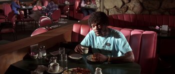 Movie still from “Pulp Fiction” (1994), directed by Quentin Tarantino – A man sitting at a table with a plate of food and a cup; Medium shot, Low angle
