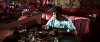 Movie still from “Pulp Fiction” (1994), directed by Quentin Tarantino – A man sitting at a table eating a sandwich and drinking coffee; Medium shot, Low angle