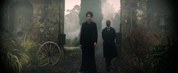 Movie still from “The Sandman” (2022), created by Neil Gaiman – A man and a man in a suit and tie standing in front of an old building; Wide shot, Low angle