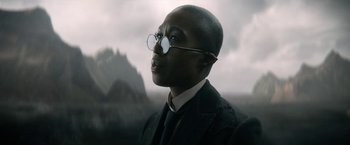 Movie still from “The Sandman” (2022), created by Neil Gaiman – A man wearing a suit and round glasses looking to his left; Close Up shot, Low angle