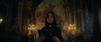 Movie still from “The Sandman” (2022), created by Neil Gaiman – A woman is holding a book in front of a painting; Medium shot, Low angle