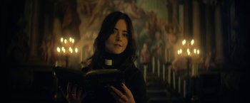 Movie still from “The Sandman” (2022), created by Neil Gaiman – A woman holding an open book in front of a painting; Close Up shot, Low angle