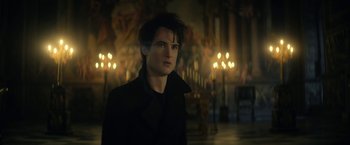 Movie still from “The Sandman” (2022), created by Neil Gaiman – A man in a black jacket standing in a room with candles; Close Up shot, Over the shoulder angle