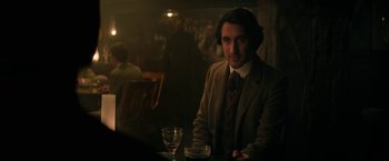 Movie still from “The Sandman” (2022), created by Neil Gaiman – A man sitting at a table with a glass of wine; Medium shot, Over the shoulder angle