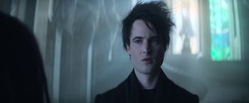 Movie still from “The Sandman” (2022), created by Neil Gaiman – A young man with dark hair and blue eyes stares at the camera; Close Up shot, Low angle