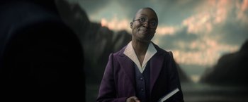 Movie still from “The Sandman” (2022), created by Neil Gaiman – A woman in a purple jacket holding an open book; Medium shot, Over the shoulder angle