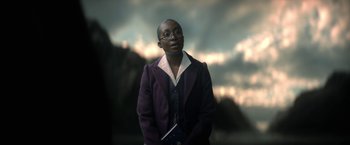 Movie still from “The Sandman” (2022), created by Neil Gaiman – A person wearing a purple jacket holding a book; Medium shot, Low angle