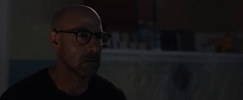 Movie still from “The Silence” (2019), directed by John R. Leonetti – A bald man with glasses is sitting in the dark; Close Up shot, Over the shoulder angle