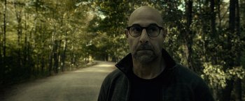 Movie still from “The Silence” (2019), directed by John R. Leonetti – A bald man wearing glasses standing on the side of a road; Close Up shot, Over the shoulder angle