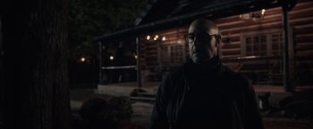 Movie still from “The Silence” (2019), directed by John R. Leonetti – A bald man wearing glasses standing in front of a log cabin; Medium shot, Over the shoulder angle