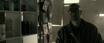 Movie still from “The Silence” (2019), directed by John R. Leonetti – A man with glasses is standing in front of a rack of books; Close Up shot, Low angle