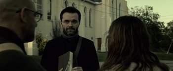 Movie still from “The Silence” (2019), directed by John R. Leonetti – A man with a beard is holding a book and talking to a woman in front of a building; Medium shot, Low angle