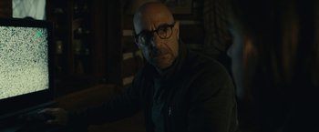 Movie still from “The Silence” (2019), directed by John R. Leonetti – A bald man with glasses is sitting in a dark room; Close Up shot, Over the shoulder angle