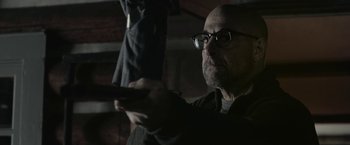 Movie still from “The Silence” (2019), directed by John R. Leonetti – A bald man with glasses is holding a remote control; Close Up shot, Over the shoulder angle